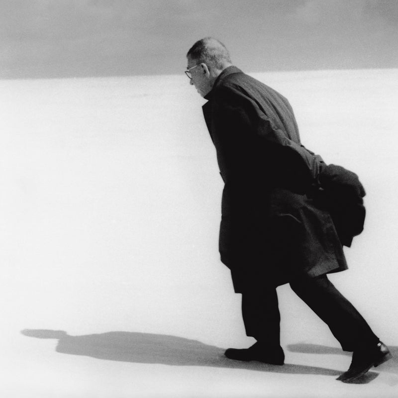 Black and white image of Sartre all dressed in black walking on a white beach against a grey sky. His shadow against the sand. Photograph by Antanas Sutkus, available as fine art print