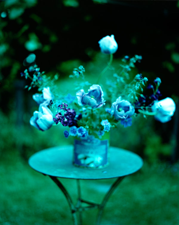 A bouquet of white peonies and blue flowers in a glass vase, on a round garden table. The background is green and black. 