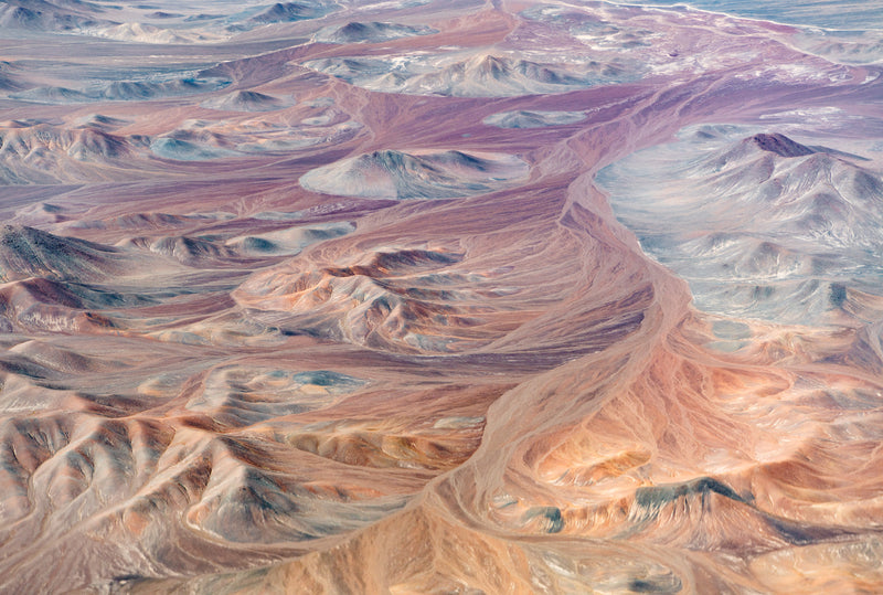 Colour photograph of the Atacama desert, shot from above, by Rowan Thornhill. Available as poster. A beautiful landscape in muted tones of blue, purple, red and orange. The landscape of mountains and valleys swirl into the horizon, making the image into abstract art.